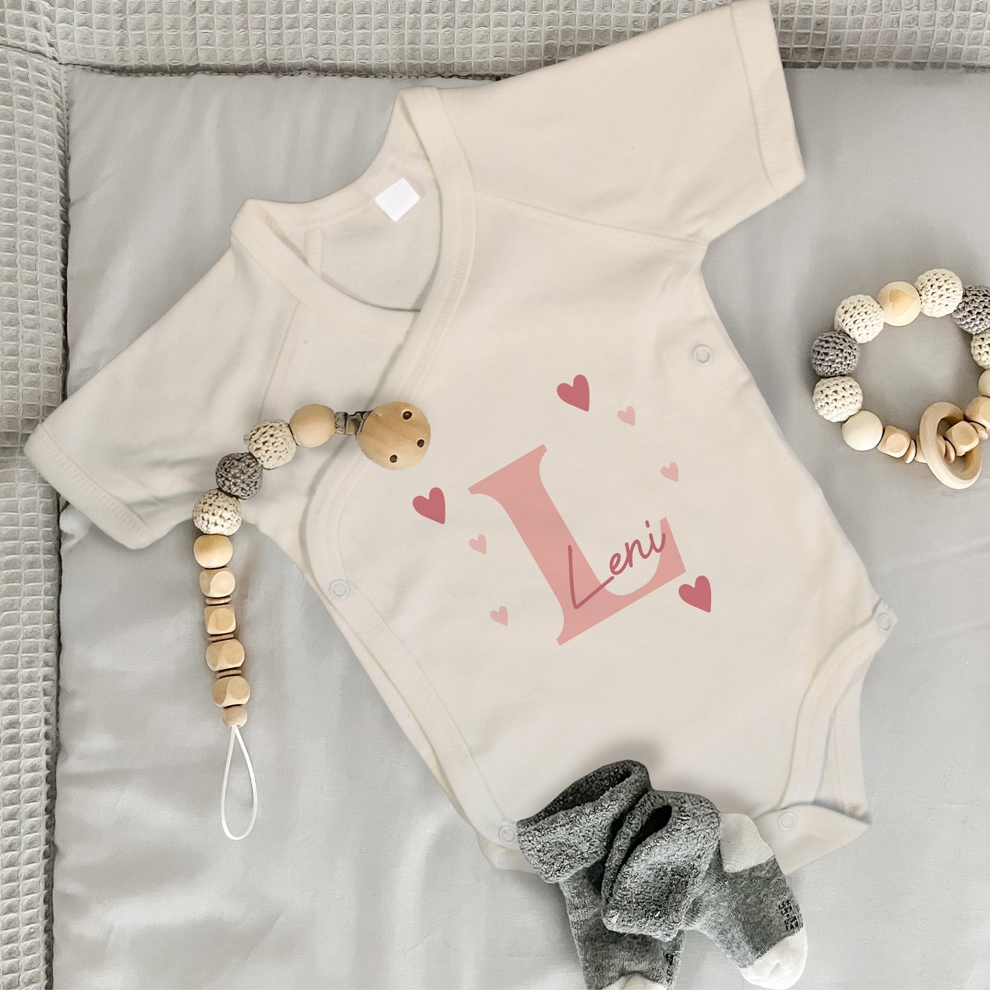 Babybody in natur mit rosa Initiale „L“, Name „Leni“ und Herzmotiven, daneben Babysocken, Beißring und Schnullerkette.