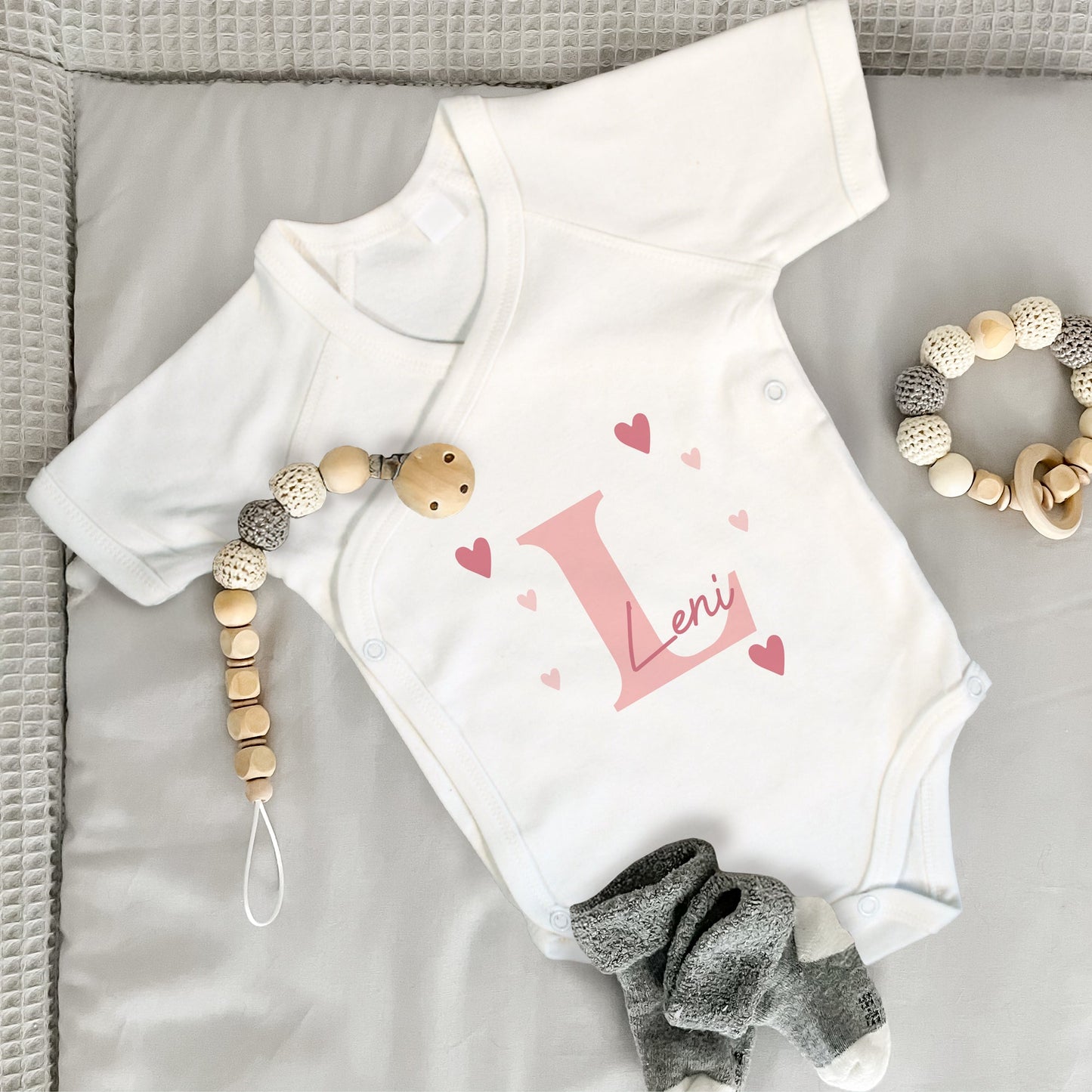 Weißer Babybody mit rosa Initiale „L“, Name „Leni“ und Herzmotiven, daneben Babysocken, Beißring und Schnullerkette.