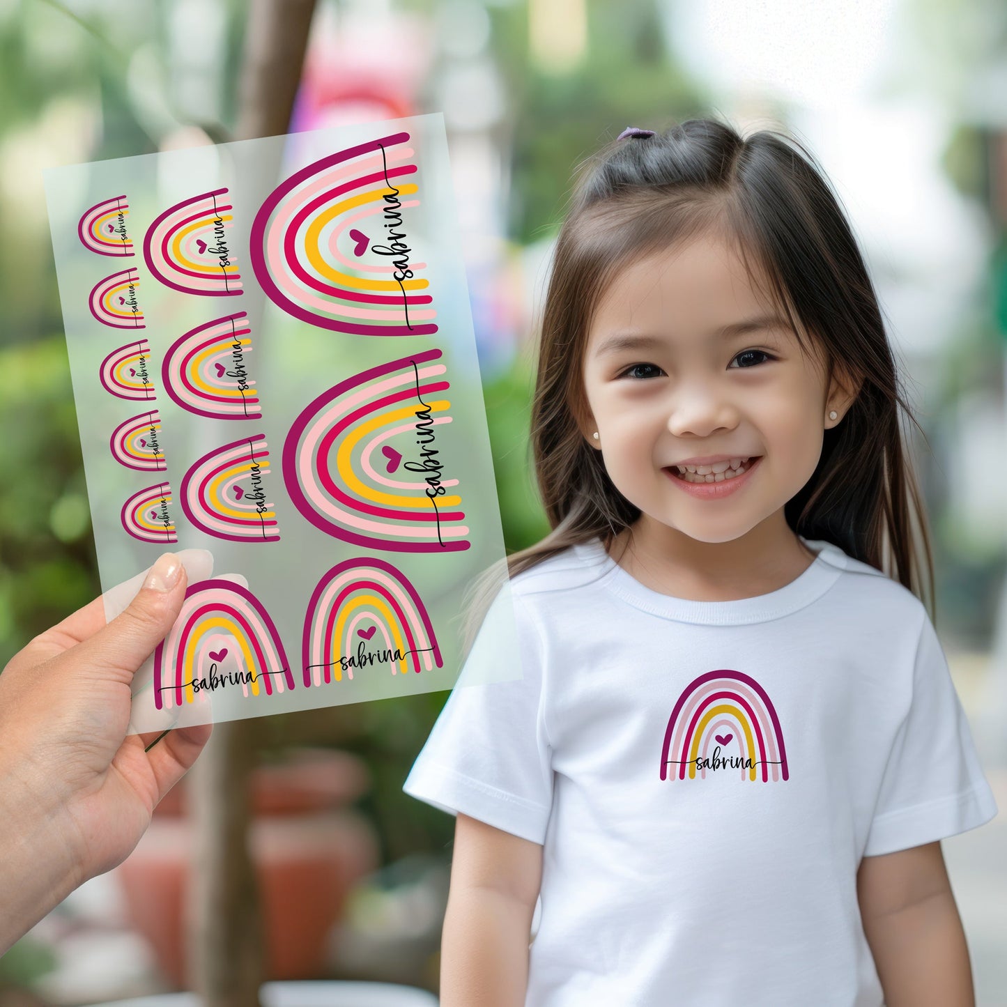 Kind trägt weißes T-Shirt mit aufgebügeltem Regenbogen-Motiv und personalisiertem Namen „sabrina“, Bügelbild-Bogen wird im Vordergrund gezeigt