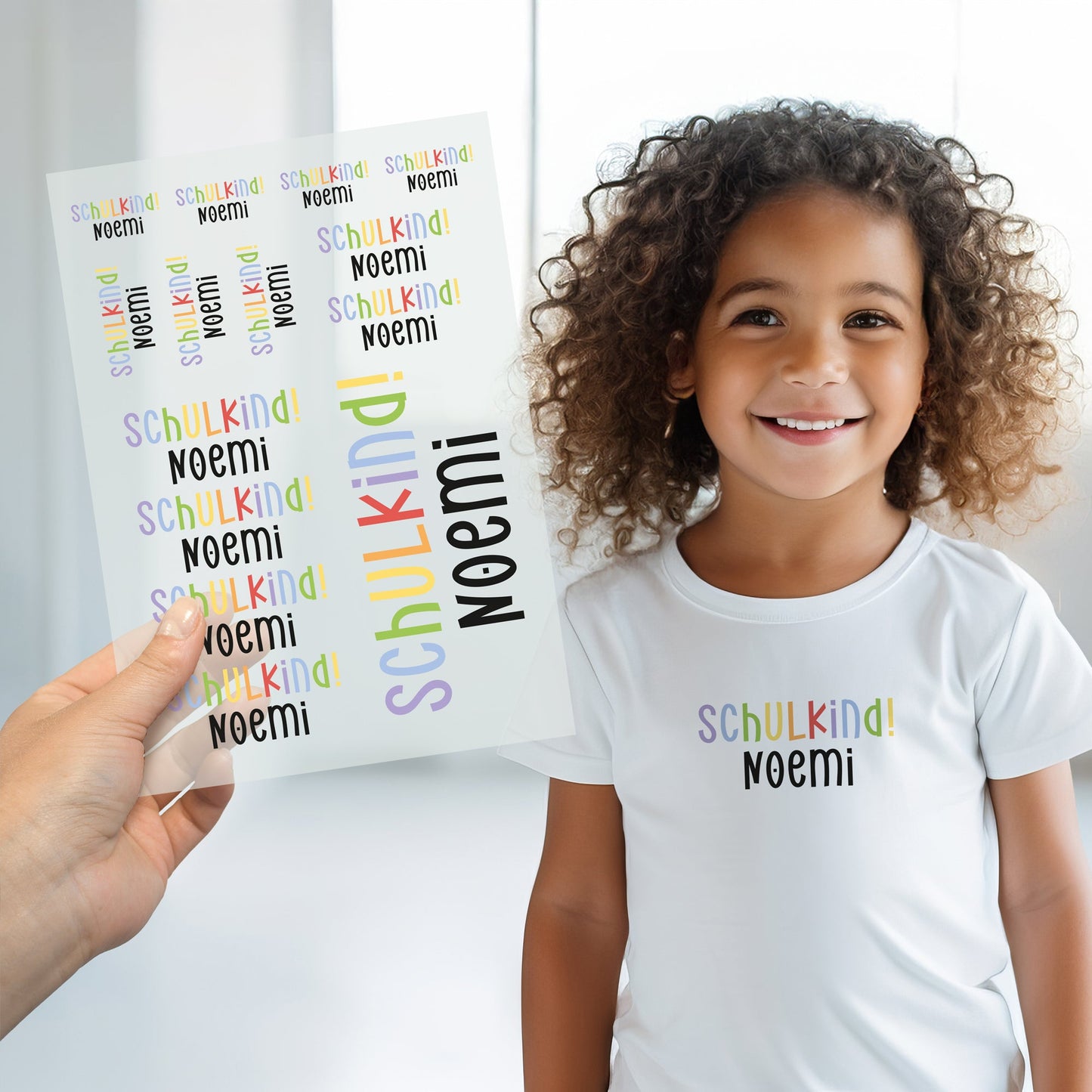 Kind trägt weißes T-Shirt mit aufgebügeltem Schulkind-Motiv, darunter der personalisierte Name „Noemi“, Bügelbild-Bogen wird im Vordergrund gezeigt
