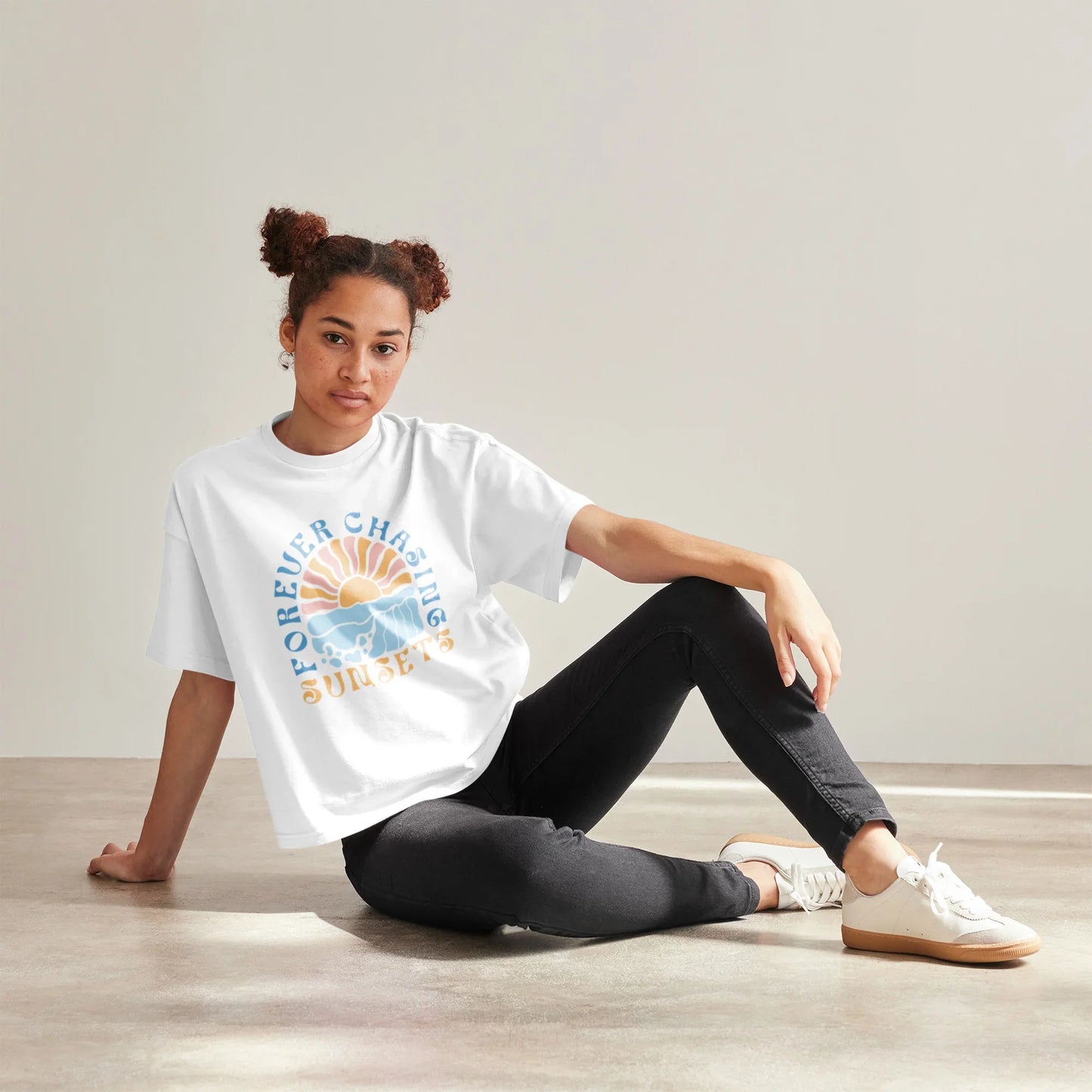 Frau trägt weißes T-Shirt mit Aufdruck 'Forever Chasing Sunsets', sitzt entspannt auf Boden.