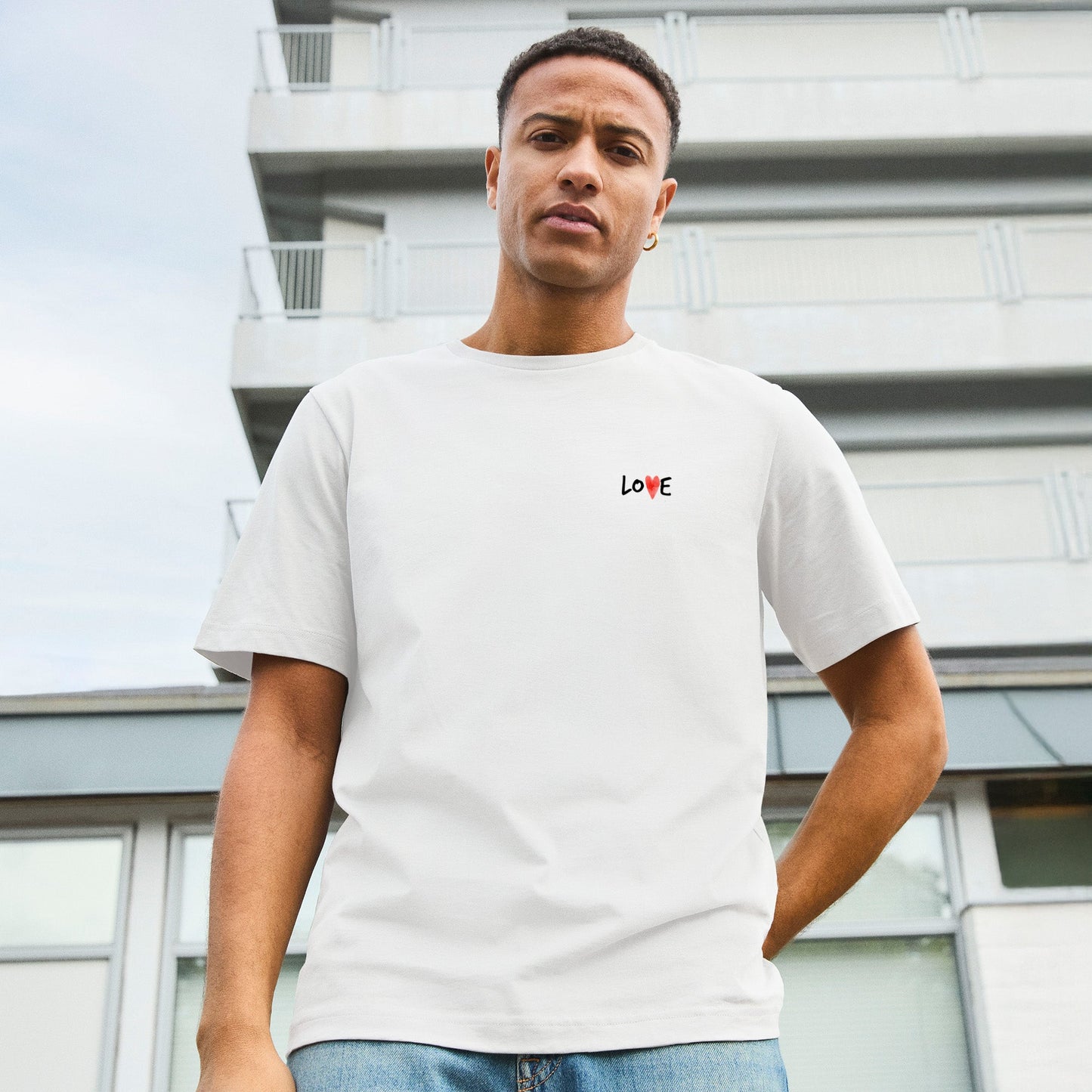 Modell trägt weißes T-Shirt mit kleinem „LOVE“-Schriftzug auf Brusthöhe, das „O“ ersetzt durch ein rotes Herz. Auf der Rückseite großer Print „LOVE IS LOVE“ mit Regenbogenherzen - als Symbol für Stolz, Akzeptanz und die LGBTQIA+ Community.