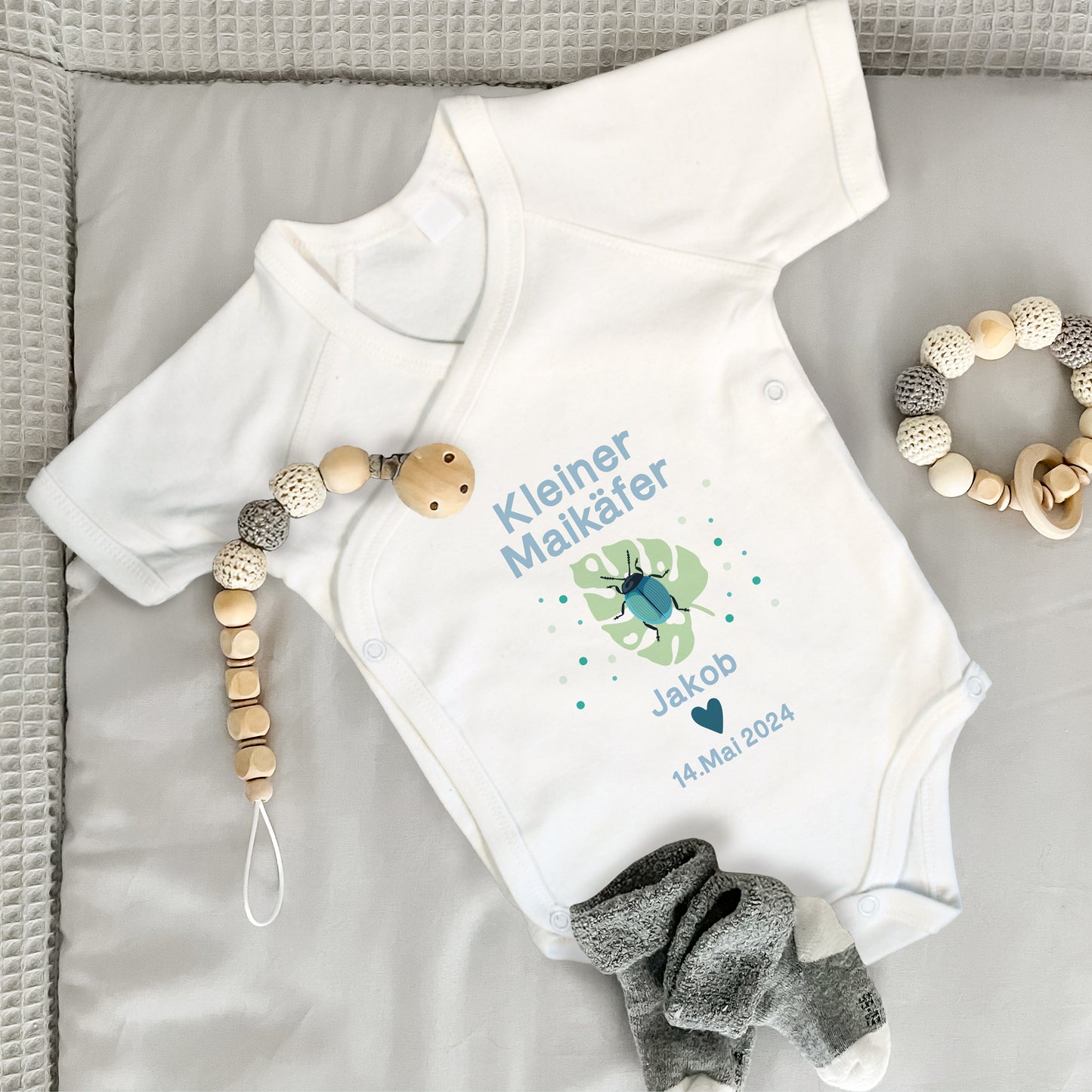 Baby Wickelbody weiß, Kurzarm auf grauer Wickelunterlage, mit Kleiner Maikäfer in Bild und blauer Schrift, mit "Jakob" und "14. Mai. 2024" bedruckt