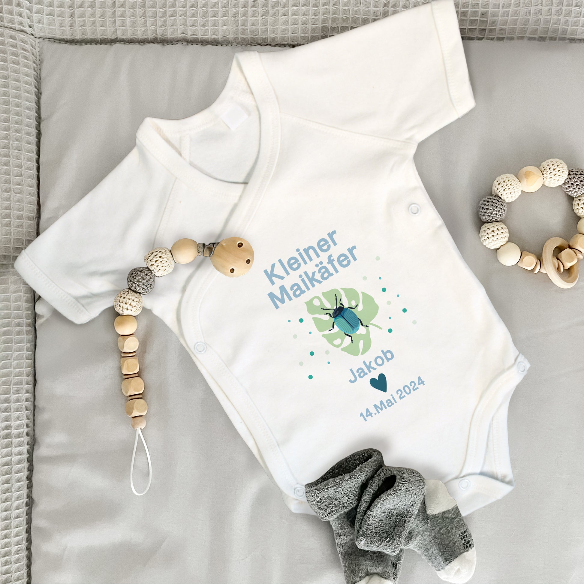 Baby Wickelbody weiß, Kurzarm auf grauer Wickelunterlage, mit Kleiner Maikäfer in Bild und blauer Schrift, mit "Jakob" und "14. Mai. 2024" bedruckt