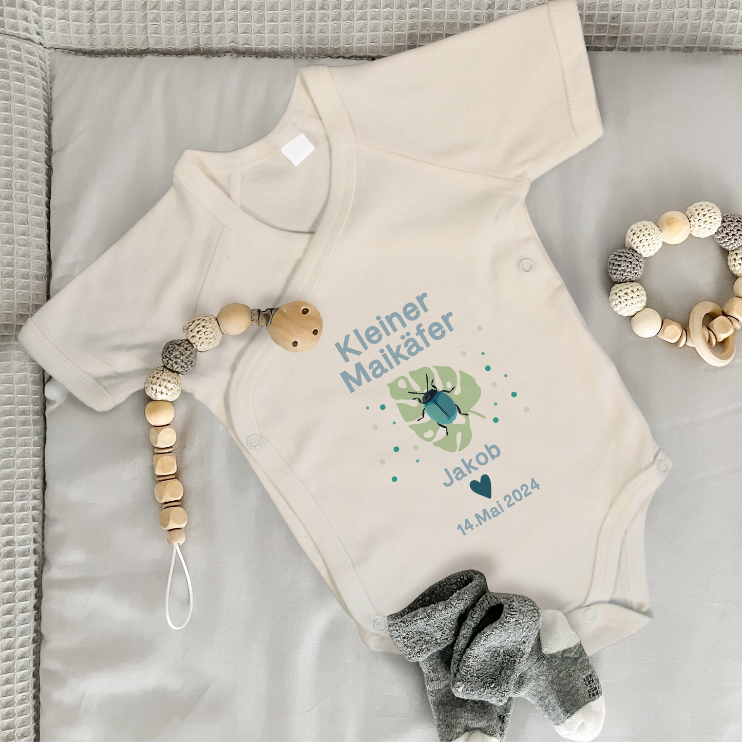 Baby Wickelbody natur, Kurzarm auf grauer Wickelunterlage, mit Kleiner Maikäfer in Bild und blauer Schrift, mit "Jakob" und "14. Mai. 2024" bedruckt