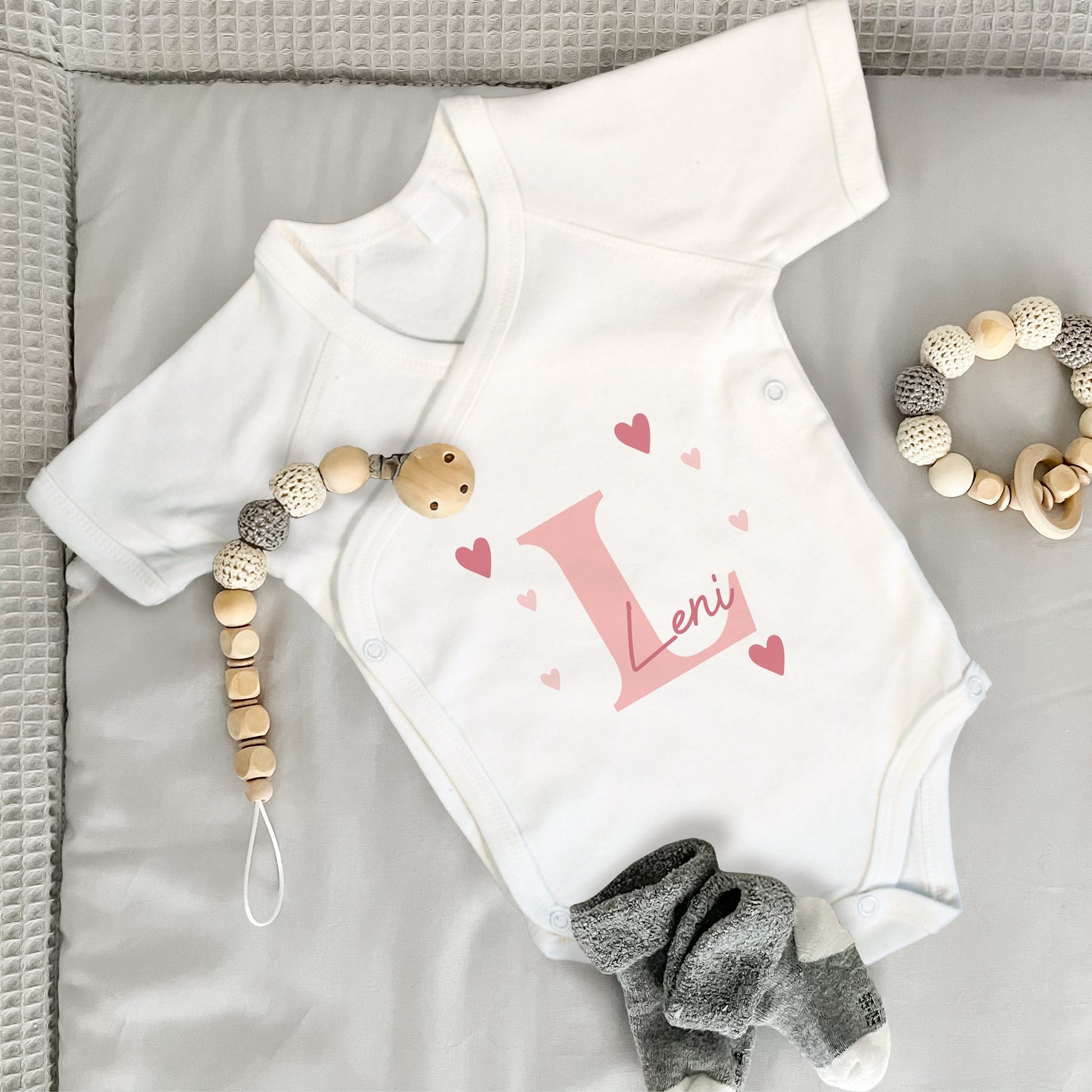 Weißer Babybody mit rosa Initiale „L“, Name „Leni“ und Herzmotiven, daneben Babysocken, Beißring und Schnullerkette.