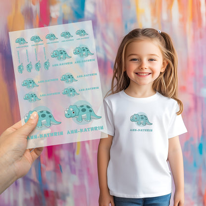 Kind trägt weißes T-Shirt mit aufgebügeltem Dino-Motiv und personalisiertem Namen „Ann-Kathrin“, Bügelbild-Bogen wird im Vordergrund gezeigt