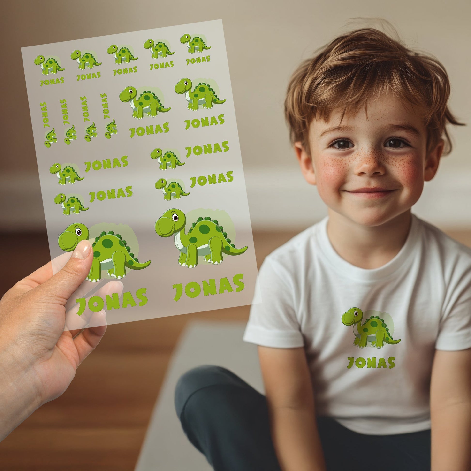 Kind trägt weißes T-Shirt mit aufgebügeltem Dino-Motiv und personalisiertem Namen „Jonas“, Bügelbild-Bogen wird im Vordergrund gezeigt