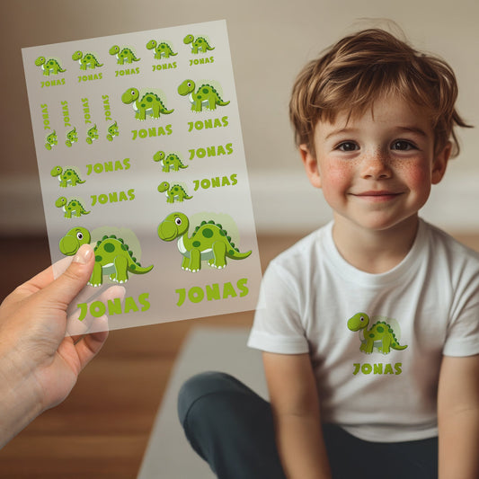 Kind trägt weißes T-Shirt mit aufgebügeltem Dino-Motiv und personalisiertem Namen „Jonas“, Bügelbild-Bogen wird im Vordergrund gezeigt