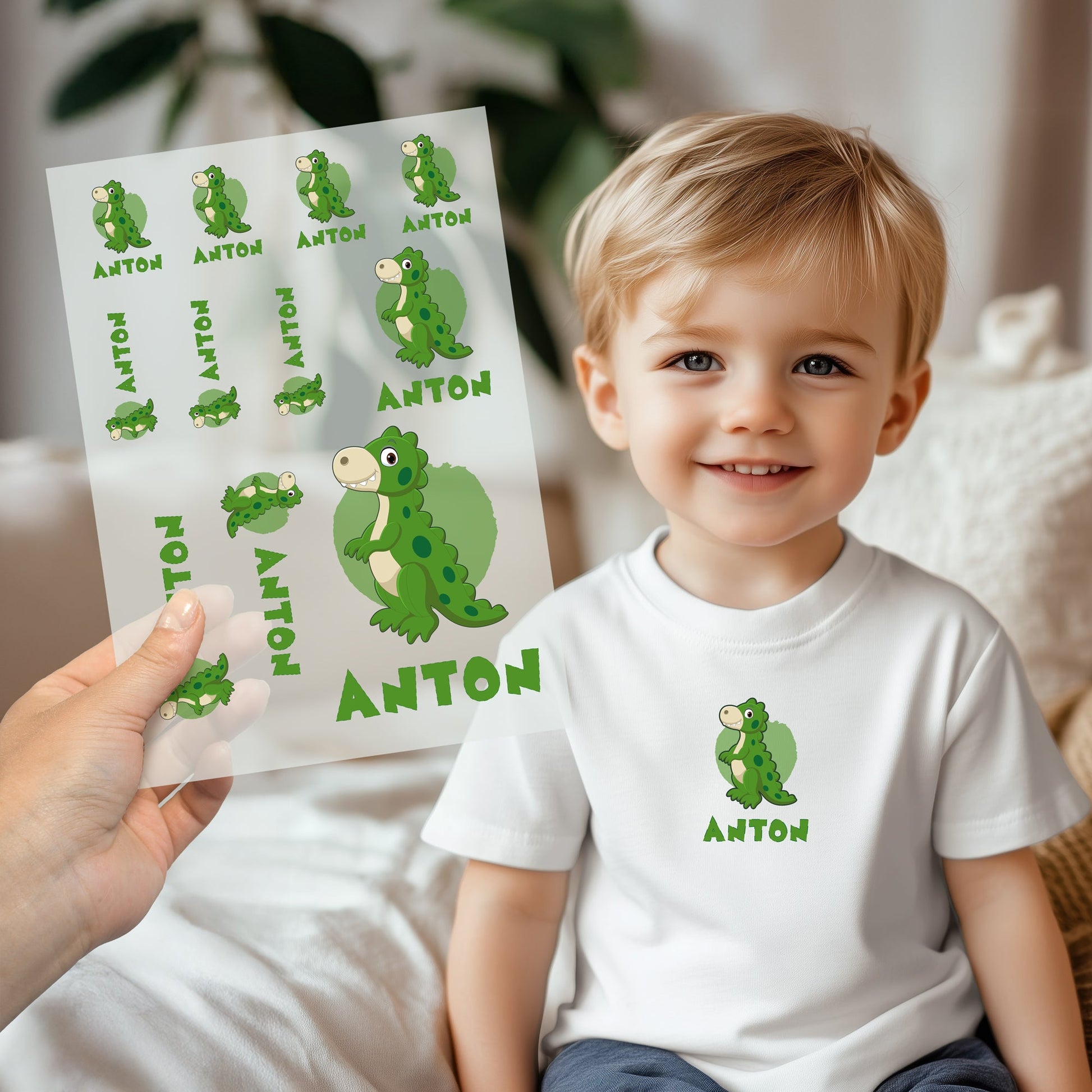 Kind trägt weißes T-Shirt mit aufgebügeltem Dino-Motiv und personalisiertem Namen „Anton“, Bügelbild-Bogen wird im Vordergrund gezeigt