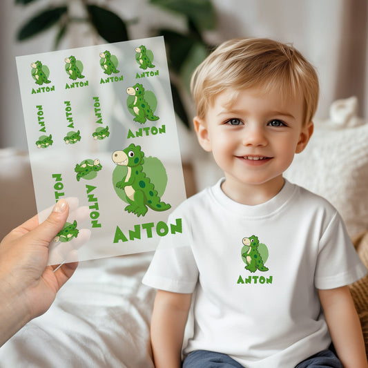 Kind trägt weißes T-Shirt mit aufgebügeltem Dino-Motiv und personalisiertem Namen „Anton“, Bügelbild-Bogen wird im Vordergrund gezeigt