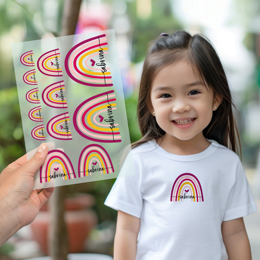 Kind trägt weißes T-Shirt mit aufgebügeltem Regenbogen-Motiv und personalisiertem Namen „sabrina“, Bügelbild-Bogen wird im Vordergrund gezeigt