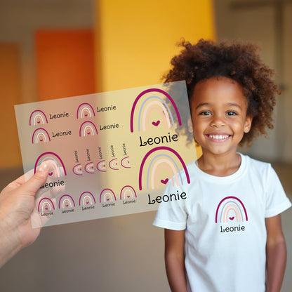 Kind trägt weißes T-Shirt mit aufgebügeltem Regenbogen-Motiv und personalisiertem Namen „Leonie“, Bügelbild-Bogen wird im Vordergrund gezeigt