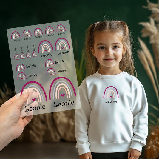 Kind trägt weißen Pullover mit aufgebügeltem Regenbogen-Motiv und personalisiertem Namen „Leonie“, Bügelbild-Bogen wird im Vordergrund gezeigt
