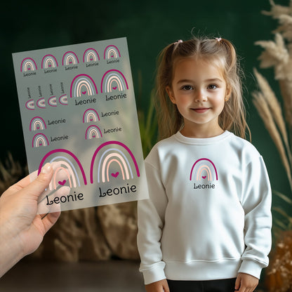 Kind trägt weißen Pullover mit aufgebügeltem Regenbogen-Motiv und personalisiertem Namen „Leonie“, Bügelbild-Bogen wird im Vordergrund gezeigt