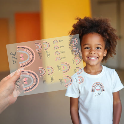 Kind trägt weißes T-Shirt mit aufgebügeltem Regenbogen-Motiv und personalisiertem Namen „Julia“, Bügelbild-Bogen wird im Vordergrund gezeigt