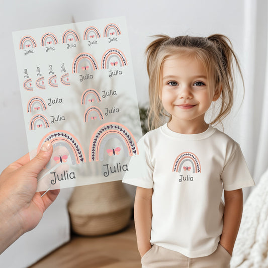 Kind trägt weißes T-Shirt mit aufgebügeltem Regenbogen-Motiv und personalisiertem Namen „Julia“, Bügelbild-Bogen wird im Vordergrund gezeigt