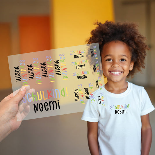 Kind trägt weißes T-Shirt mit aufgebügeltem Schulkind-Motiv, darunter der personalisierte Name „Noemi“, Bügelbild-Bogen wird im Vordergrund gezeigt