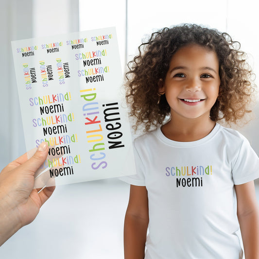 Kind trägt weißes T-Shirt mit aufgebügeltem Schulkind-Motiv, darunter der personalisierte Name „Noemi“, Bügelbild-Bogen wird im Vordergrund gezeigt