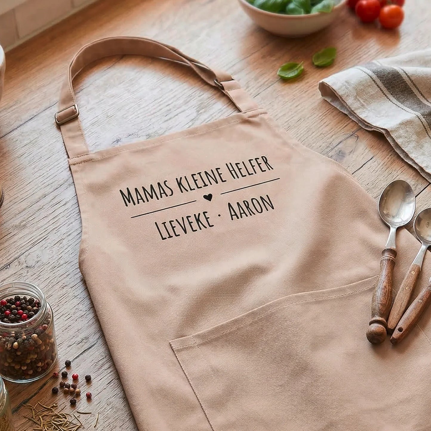 Personalisierte Schürze in Beige mit Aufschrift, auf Holztisch mit Küchenutensilien dekoriert.