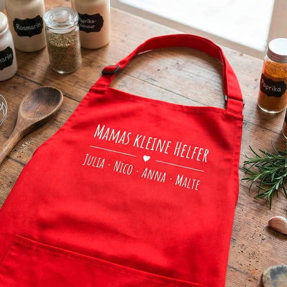 Rote personalisierte Schürze mit Namen, Küchenutensilien und Kräutern auf Holztisch
