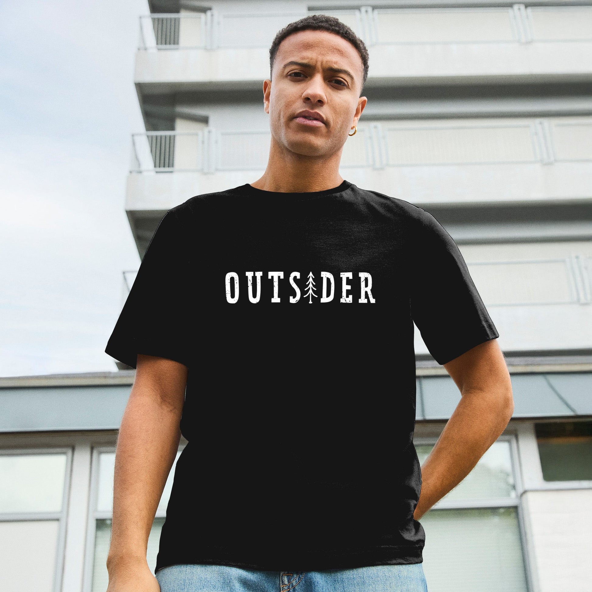 Modell trägt schwarzes T-Shirt mit Frontdruck „OUTSIDER“, bei dem das „i“ durch eine stilisierte Tanne ersetzt ist